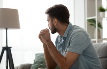 A man in a blue shirt sits on a couch, looking pensively out a window.